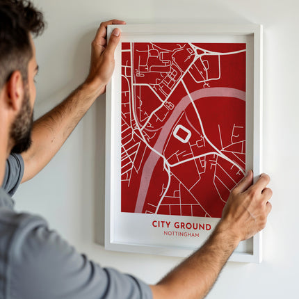 A man hanging a framed map print, featuring a contemporary style illustration of the City Ground, home to Nottingham.