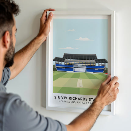 A man hanging a framed art print, featuring a contemporary style illustration of the Sir Viv Richards Stadium, home to the West Indies and Antigua & Barbuda.